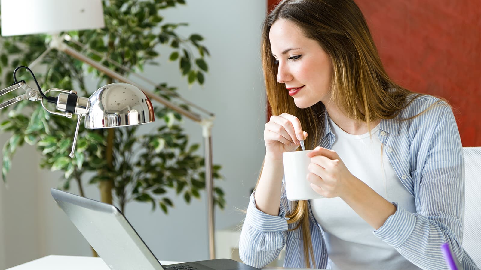 woman stirring coffee in her cup while looking at a laptop