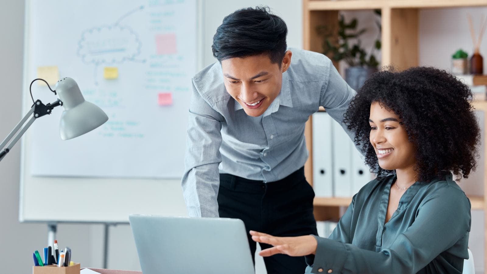 Woman seated, man standing, looking at same laptop
