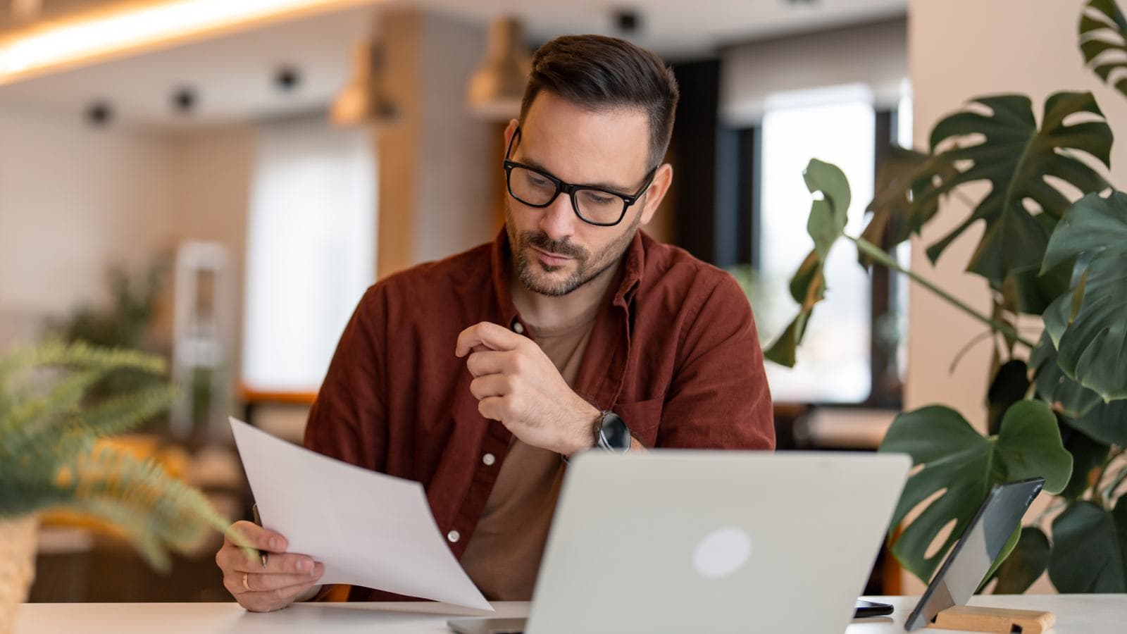 Man perusing document next to big plant and laptop