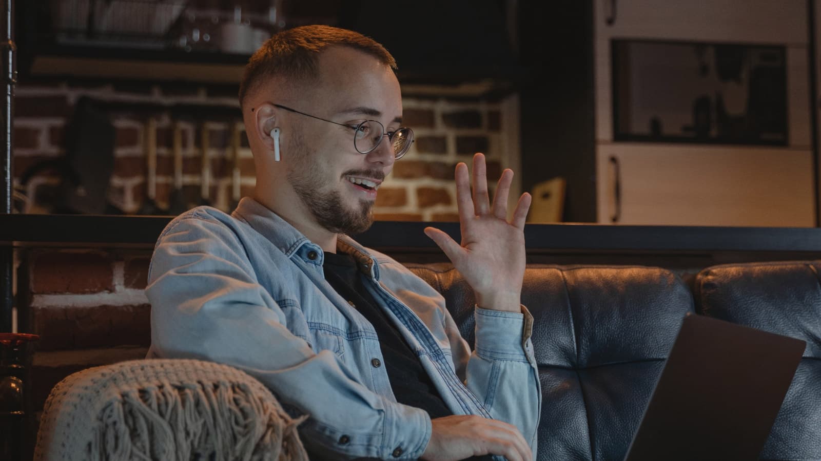 Man on sofa wearing earbuds, waving at laptop